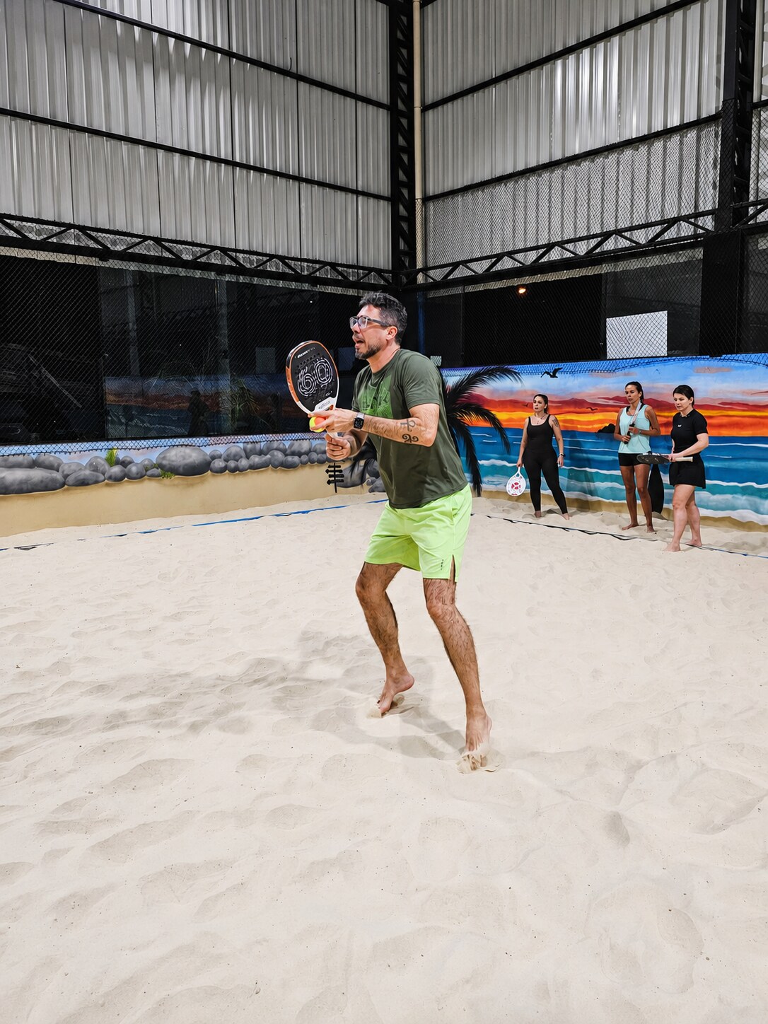 Jogador correndo na areia para devolver a bola, com o mural de praia ao fundo